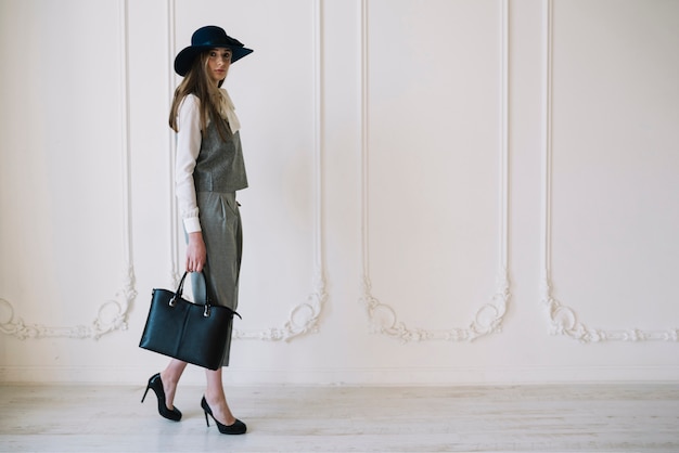 Stylish young woman in costume and hat with handbag in room