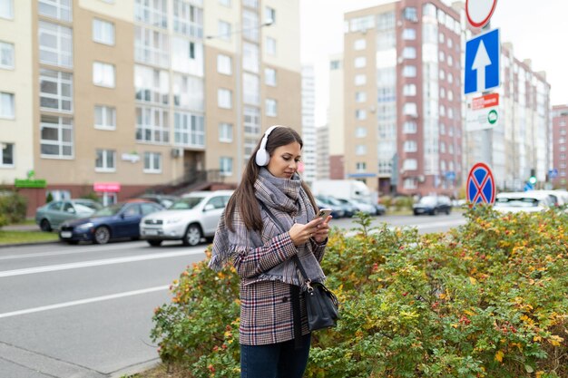 ヘッドフォンで横断歩道を歩くスタイリッシュな若い女性が携帯電話を見る