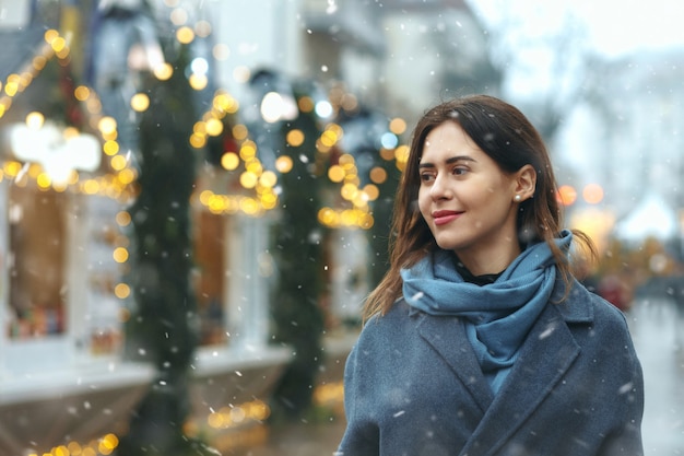 Stylish young woman dressed in trendy coat walking at the street during the snowfall. Empty space