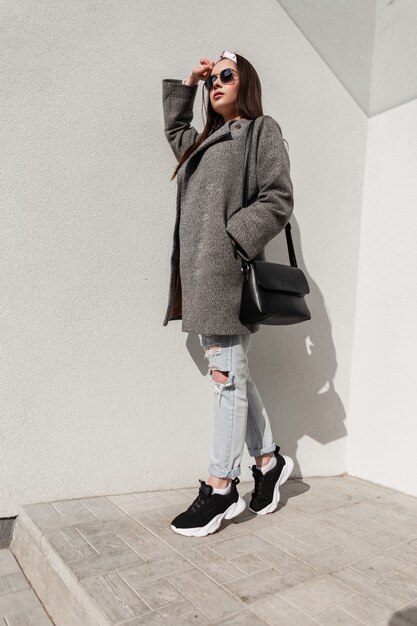 Stylish young woman in fashionable casual youth coat with handbag in sneakers with bandana in sunglasses is resting on sunny day near vintage wall on street. Attractive urban girl and bright sun.