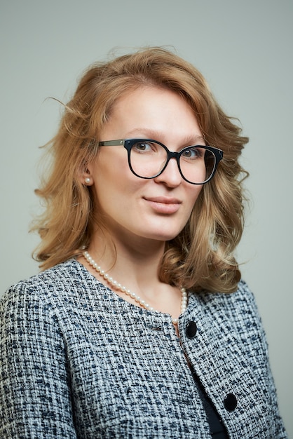 A stylish young woman in glasses dressed in a grey skirt suit with a black shirt. A close-up portrait of a blond lady who wears a business outfit.