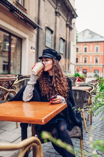 Foto elegante giovane donna che mangia caffè nella caffetteria all'aperto mentre si utilizza lo smartphone. ritratto di moda di bellezza di una ragazza del college che si rilassa sulla strada della città