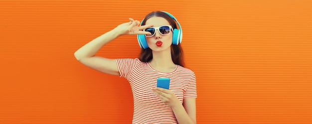 オレンジ色の背景でスマートフォンで音楽を聴いているヘッドフォンをかぶったスタイリッシュな若い女性