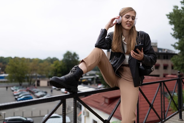 Стильная молодая женщина в наушниках слушает музыку с мобильного телефона, положив ногу на парапет