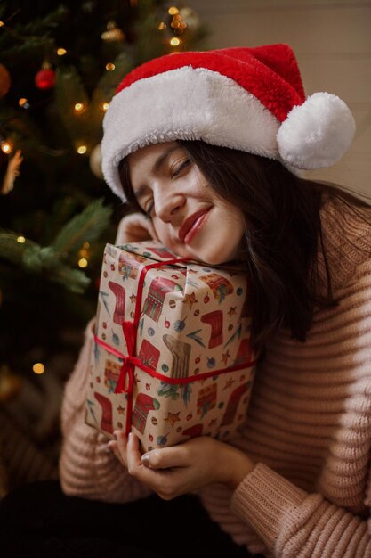 Stylish young woman holding christmas gift box and dreaming under christmas tree with lights in modern room Happy girl in santa hat receiving christmas present Happy holidays