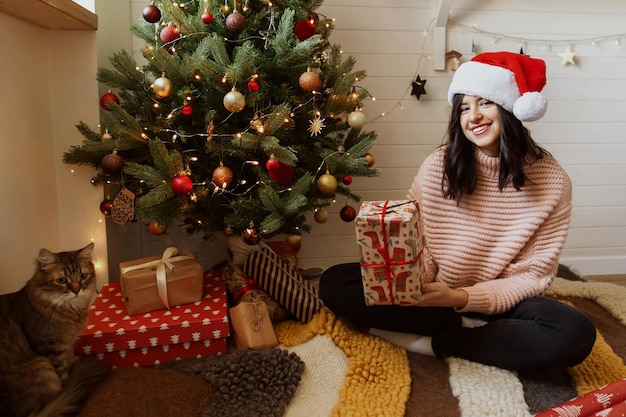 Stylish young woman holding red christmas gift box under\
christmas tree with lights in modern room happy girl in santa hat\
and sweater preparing christmas presents happy holidays