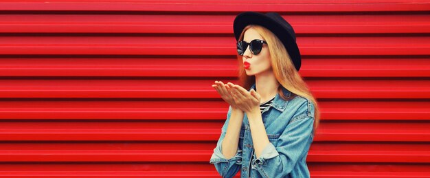 Stylish young woman model blowing kiss in jean jacket black round hat on red background