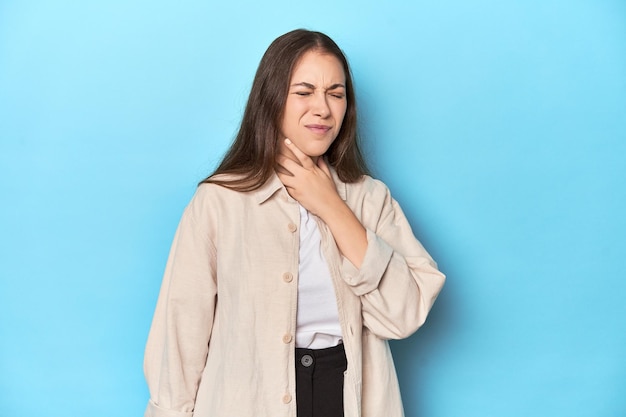 Stylish young woman in an overshirt on a blue background suffers pain in throat due a virus
