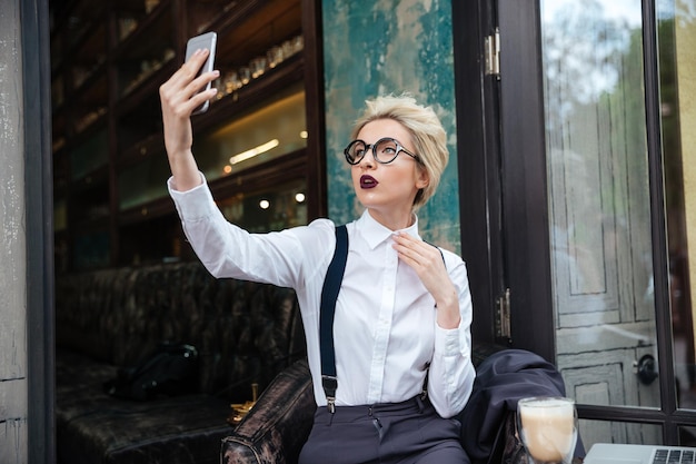 Stylish young woman in round glasses taking selfie in cafe