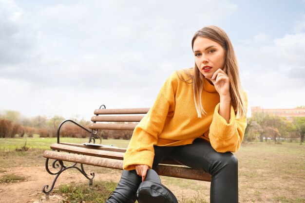 Stylish young woman sitting on bench in park on autumn day