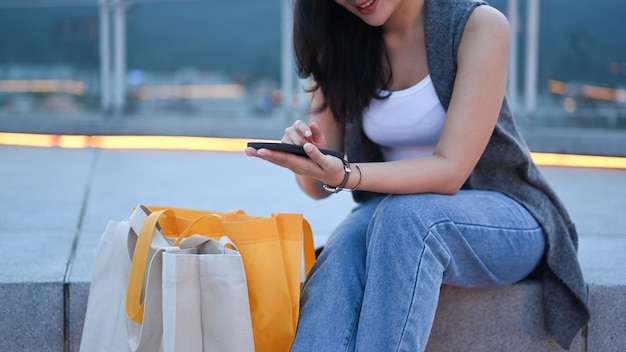 Stylish young woman sitting on stairs with shopping bags and using smart phone resting after shopping