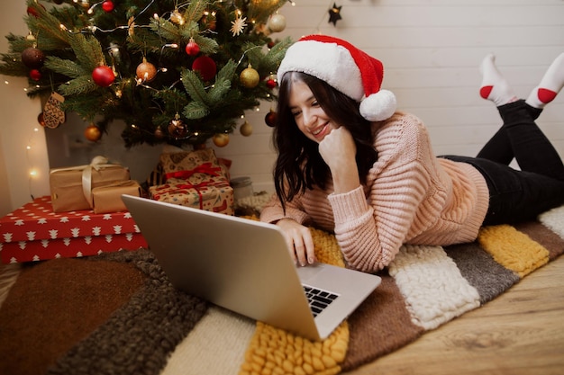 Stylish young woman sitting with laptop on rug under christmas tree with presents in modern room Happy girl in santa hat working or shopping on christmas sales Happy holidays