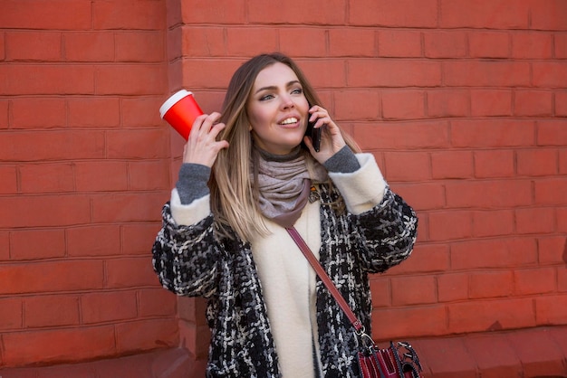 Elegante giovane donna che parla al telefono in città con caffè artigianale in mano sullo sfondo di un edificio in mattoni rossi emozioni positive camminano nella città autunnale