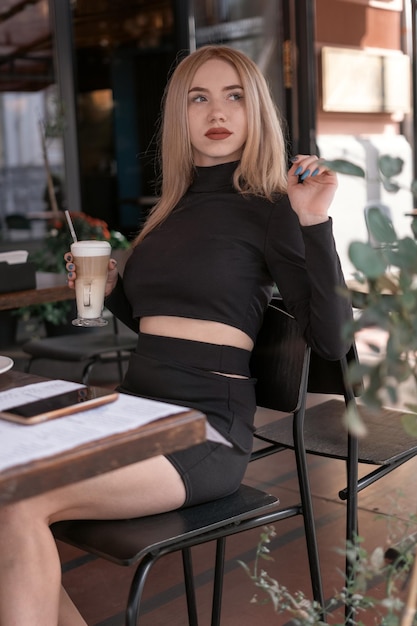 Stylish young woman in tight black clothes sits at table in cafe and holds latte or cappuccino coffee Vertical frame