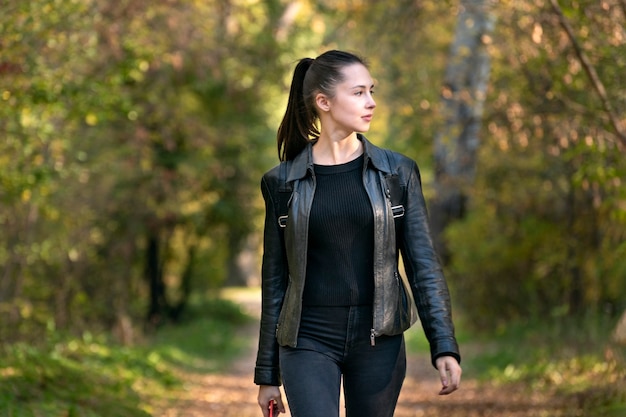 Stylish young woman walks in the autumn park. Beautiful girl in black clothes in the forest. Alley, promenade.