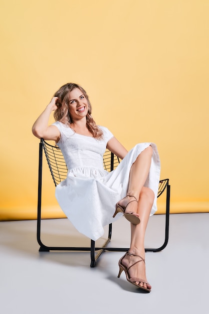 Stylish young woman in white sundress sitting in metal armchair in studio