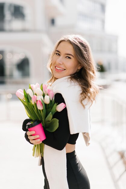 Фото Стильная молодая женщина с букетом тюльпанов на открытом воздухе и держащая бумажную чашку с кофе в руках портрет весеннего уличного образа жизни