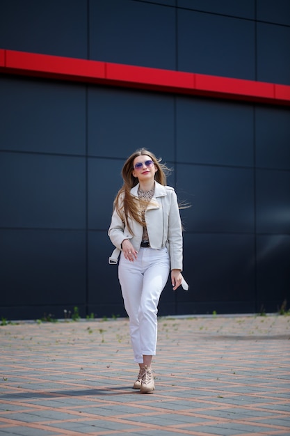 Stylish young woman with long blond hair of European appearance with a smile on her face. Girl in a white jacket and white jeans a warm summer sunny day on a background of a gray building