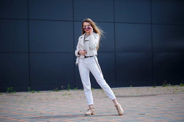 Stylish young woman with long blond hair of European appearance with a smile on her face. Girl in a white jacket and white jeans a warm summer sunny day on a background of a gray building