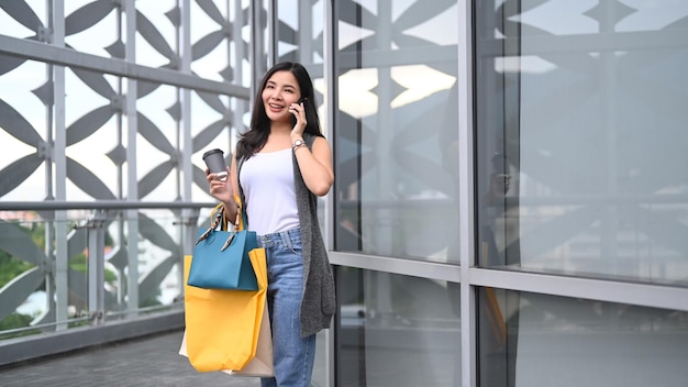 Giovane donna alla moda con le borse della spesa e parlando al cellulare che cammina fuori dal centro commerciale