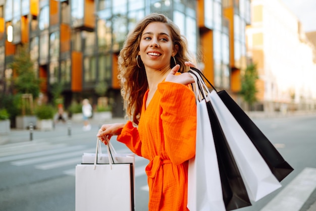 Giovane donna elegante con le borse della spesa che cammina per la strada soleggiata in un vestito luminoso concetto di vendite