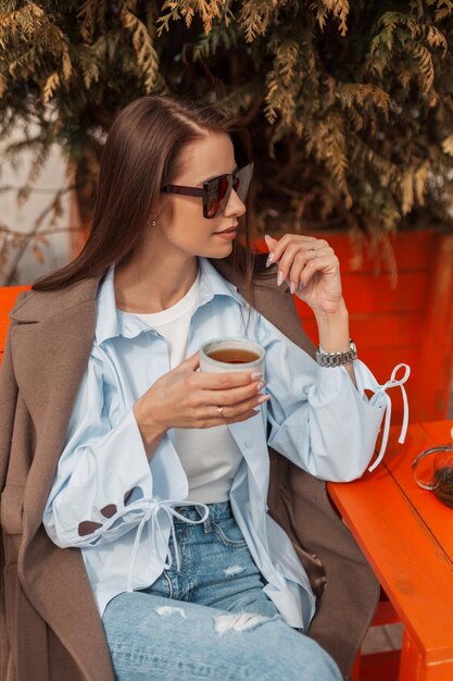 Stylish young woman with vintage sunglasses in a fashionable shirt with a coat and jeans sits on a terrace and drinks tea