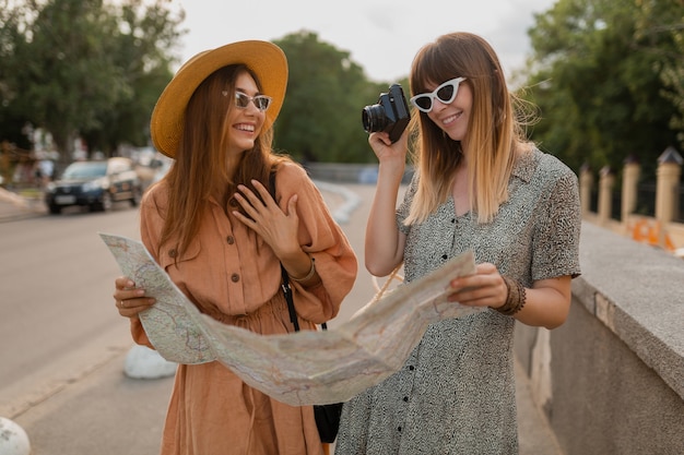 봄의 트렌디한 드레스와 액세서리를 입고 함께 여행하는 세련된 젊은 여성들이 지도를 들고 카메라에서 사진을 찍는 것을 즐겁게 하고 있습니다.