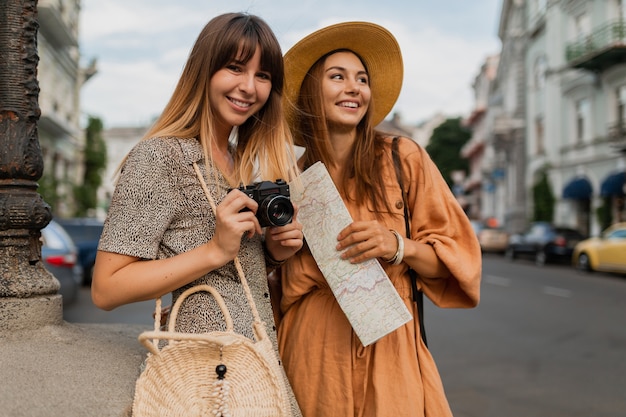 Stylish young women travelling together in Europe dressed in spring trendy dresses and accessories holding map