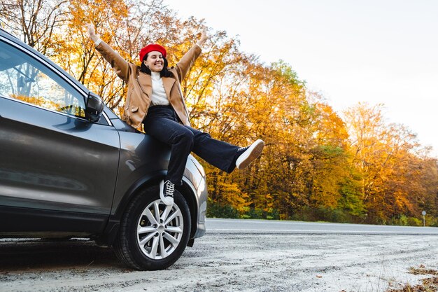 A stylishly dressed woman sits on the hood of a car with her arms raised