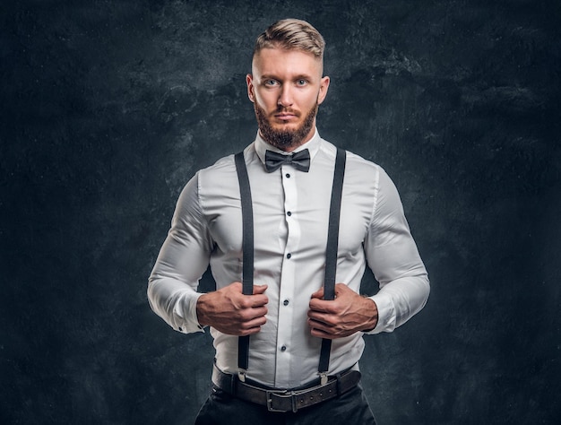 Stylishly dressed young man in shirt with bow tie and\
suspenders. studio photo against a dark wall background