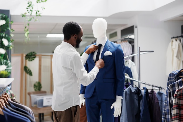 Stylist kledingwinkel die formele kleding maakt op etalagepop in winkel. Afro-amerikaanse man die dummy-model in pak aankleedt en stropdas bevestigt terwijl hij in een winkelcentrum werkt