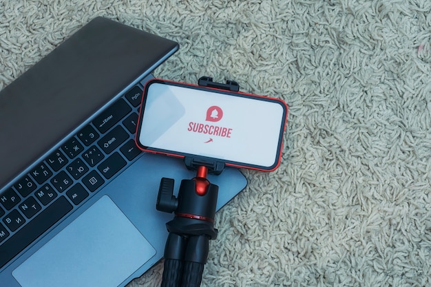 Subscribe to the Internet channel on the smartphone display on a flexible tripod with Laptop against the background of the carpet.