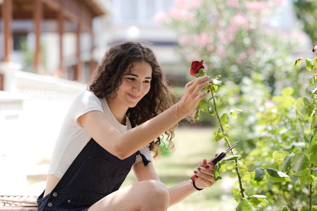 Suburban life Teen girl florist with curly hair pruning roses in the yard working in the garden Young woman with pruner cutting flowers