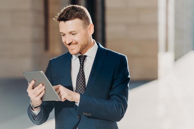 Succesful male entrepreneur in formal suit holds modern tablet\
checks documentation online reads necessary information for making\
business report uses free wifi connection people and\
technology