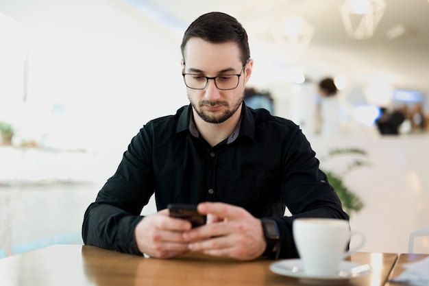 成功したリモート男性労働者。電話に満足している若いフリーランサーの男。背景のコーヒーショップ。コーヒーショップで働く男性のフリーランサープログラマー。彼はクライアントと喜んで話をします