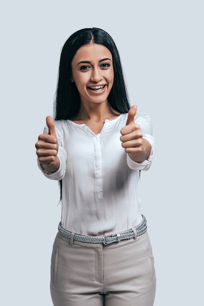 Success achieved! Happy young attractive woman in white shirt gesturing and smiling while standing against grey background