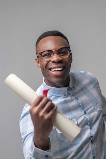 Успех, эмоции. Молодой взрослый энергичный радующийся темнокожий мужчина в очках в клетчатой рубашке с бумажным рулоном в руке