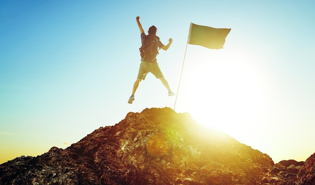 Photo success leadership achievement and people concept silhouette of young guy with flag on mountain top over sky and sun light background