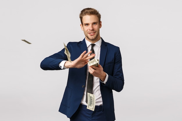 Success, money and finance concept. Handsome confident, blond bearded businessman in suit, holding cash and throwing money in air with pleased, satisfied expression, wasting dollars