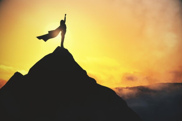 Photo success and satisfaction concept with super woman in waving coat on top of dark rock on yellow foggy sky background