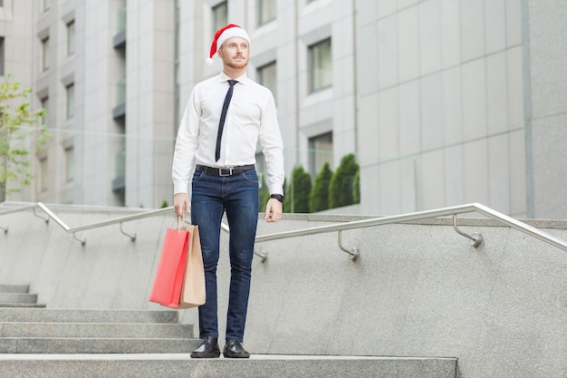 Success young adult ginger businessman with santa red hat looking away and holding gifts