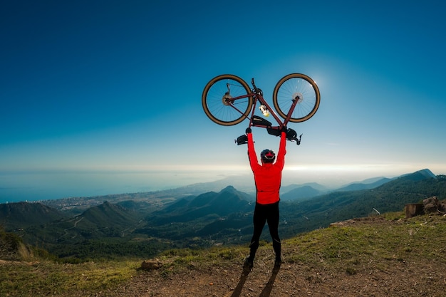 Photo successful adult male cyclist rises his by bicycle over his head on top of a mountain