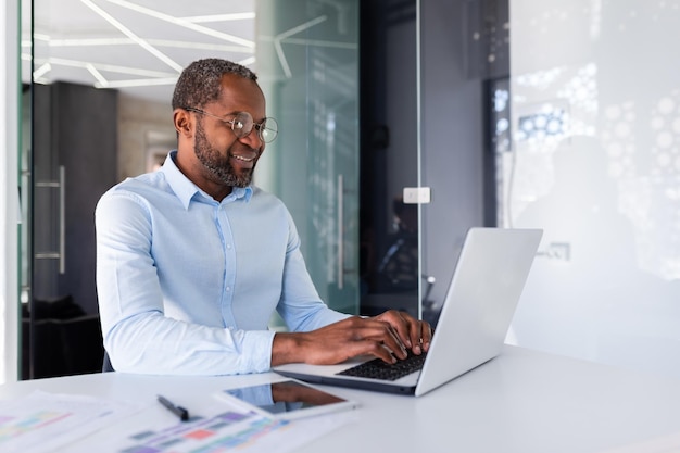 Успешный афроамериканский бизнесмен, работающий в офисе с ноутбуком, улыбающийся и довольный