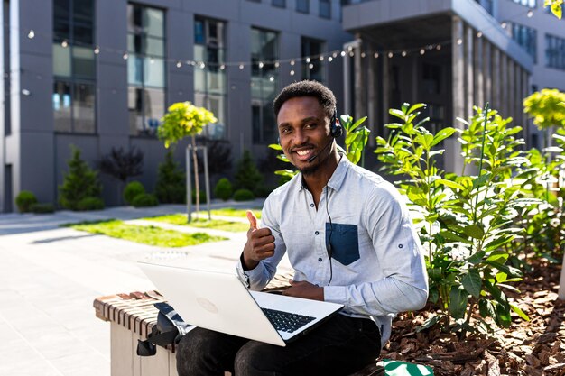 Il successo dell'uomo d'affari afroamericano lavora con il laptop e parla al telefono, guarda la telecamera e sorride gioisce all'aperto, in abiti casual