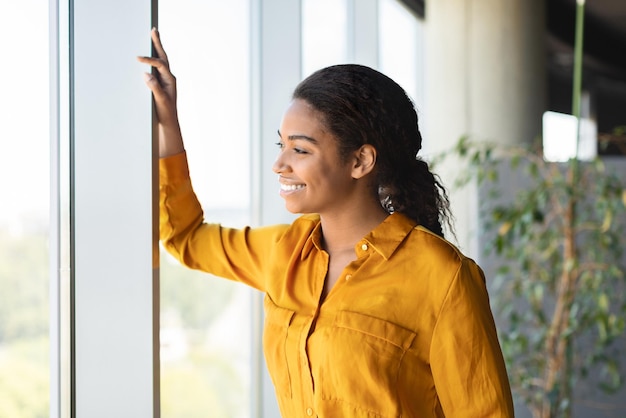 Successful african american businesswoman standing in modern coworking office and looking through window and smiling