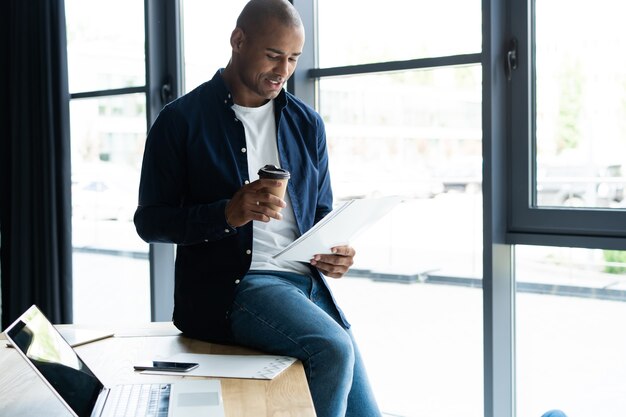 Successful African entrepreneur studying documents with attentive and concentrated look, drinking coffee at cafe. Dark-skinned businessman focused on work issues, signing papers for business deals