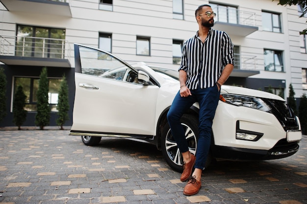 Successful arab man wear in striped shirt and sunglasses pose near his white suv car. Stylish arabian men in transport.