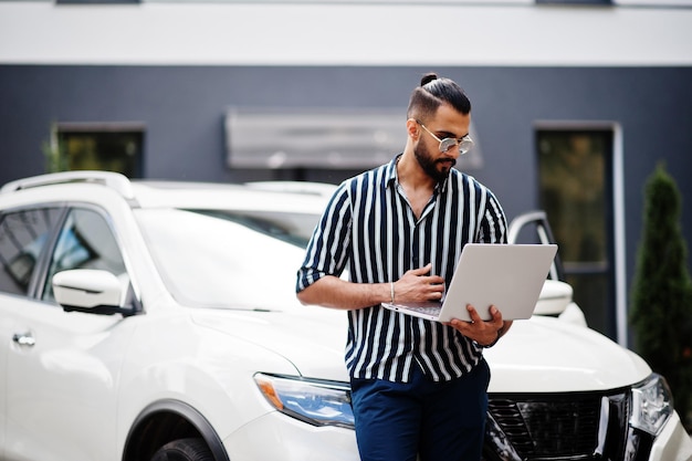 L'uomo arabo di successo indossa una camicia a righe e occhiali da sole posa vicino alla sua auto suv bianca con il laptop in mano.