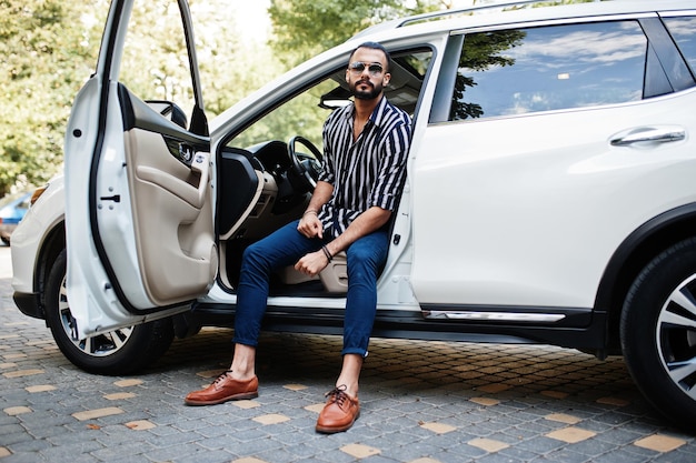 Successful arab man wear in striped shirt and sunglasses pose behind the wheel of  his white suv car. Stylish arabian men in transport.