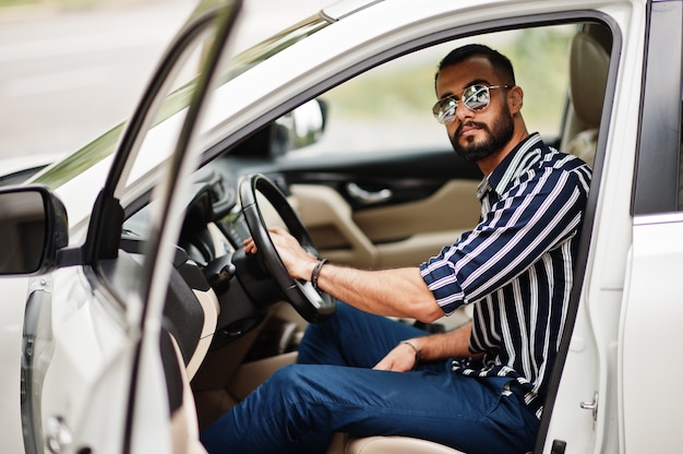 Successful arab man wear in striped shirt and sunglasses pose behind the wheel of  his white suv car. Stylish arabian men in transport.